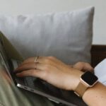 Smart Homes - Side view of crop unrecognizable female freelancer in casual clothes and smartwatch lying on comfortable sofa and working remotely on laptop