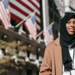 Intercultural Marriages - From below of cheerful African American female ambassador with folder wearing hijab and id card looking away while standing near building with American flags on blurred background