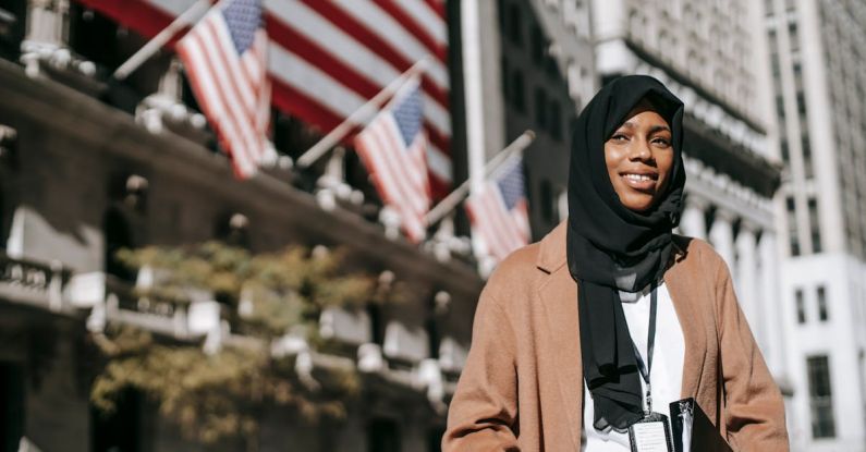 Intercultural Marriages - From below of cheerful African American female ambassador with folder wearing hijab and id card looking away while standing near building with American flags on blurred background